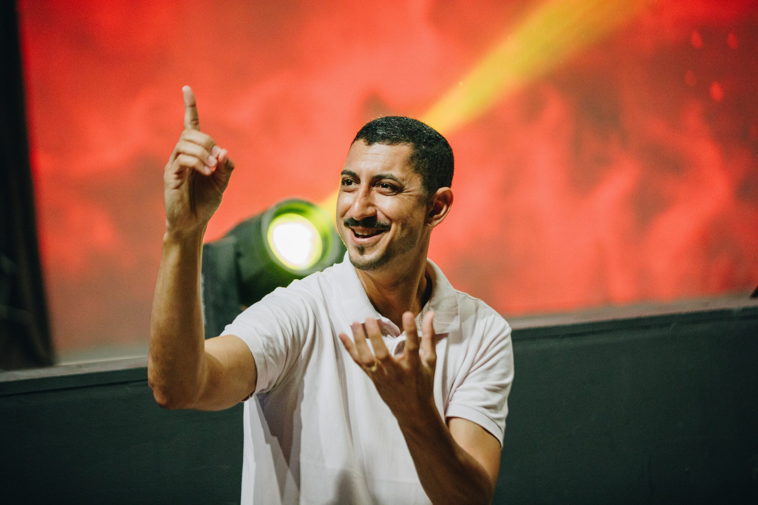 Man in white shirt standing in front of a stage pointing and using sign language