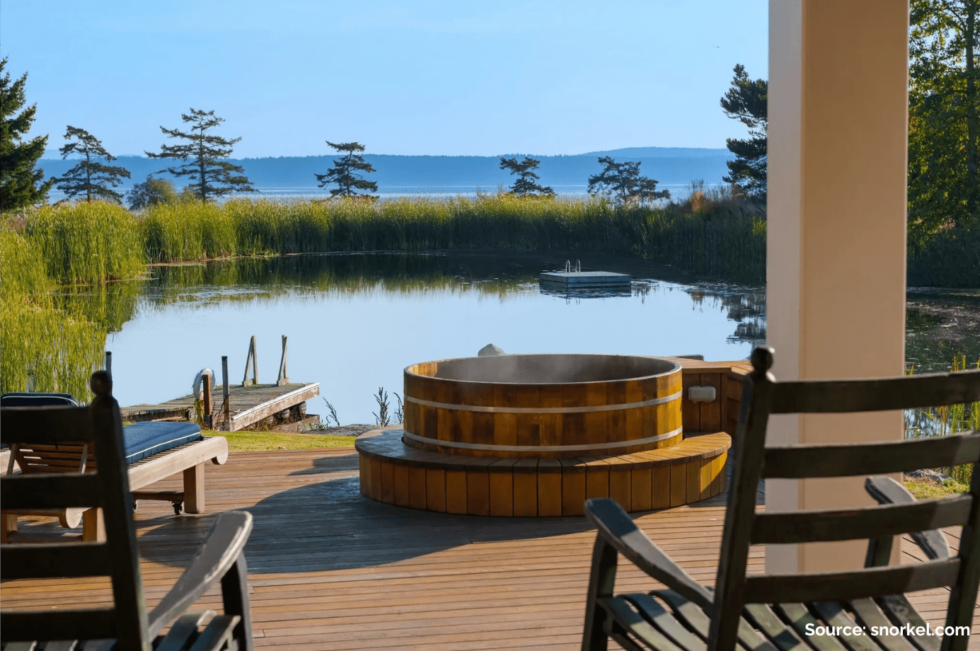 A beautiful view of rocking chairs and a wooden hot tub on a porch overlooking a pristine lake with trees and blue sky in the background